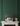 A styled interior scene with a dark green paneled wall and gold vertical lines, featuring a white marble mantel with decorative objects including a lit candle, a small framed print, a dark vase with red branches, and a gift box.