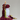 A sculptural candle shaped like a white Buddha head, with dark red wax styled like hair and dripping down the side, plus a lit flame rising from the top. Yellow-green wax accents and additional red wax pieces are arranged around the base on a plain light gray background.