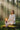 A woman in a white outfit sits cross-legged on a mossy log in a sunlit forest, holding a lit candle stack. Trees and soft light beams fill the background.