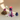 A sculptural candle with a lit flame, featuring stacked ribbed rings in pink, purple, and navy with dripping wax, displayed on a white base with decorative props against a warm beige background.