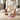 A person in a cream ribbed knit outfit sits cross-legged on a light wood floor, holding a dark blue faceted incense holder. In front of them is a white sculpted incense burner with a smoking stem and several pink silicone incense tips on a purple base.