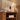 A lit table lamp with a cylindrical base and tapered cream shade sits on a wooden shelf beside stacked books and a decorative stack of round objects, against a warm wall with palm shadow cast across it.