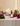 A styled product display on a white marble surface featuring a pink and purple stacked candle holder with a white sculpted face, an open white jewelry box with multiple rings, and tropical flowers and green leaves against a warm beige background.