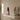 A decorative incense burner sculpture sits on a white floating shelf in a styled interior. The sculpture features a white carved face with smoke rising from its top, layered with dark red, black, and pink elements, and placed on a bright pink rectangular base.