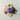 A faceted purple cosmetic jar or container arranged on a white textured surface with pink rose petals, small brown seeds, yellow flower buds, red berries, green pine needles, and a few pale stones around it.